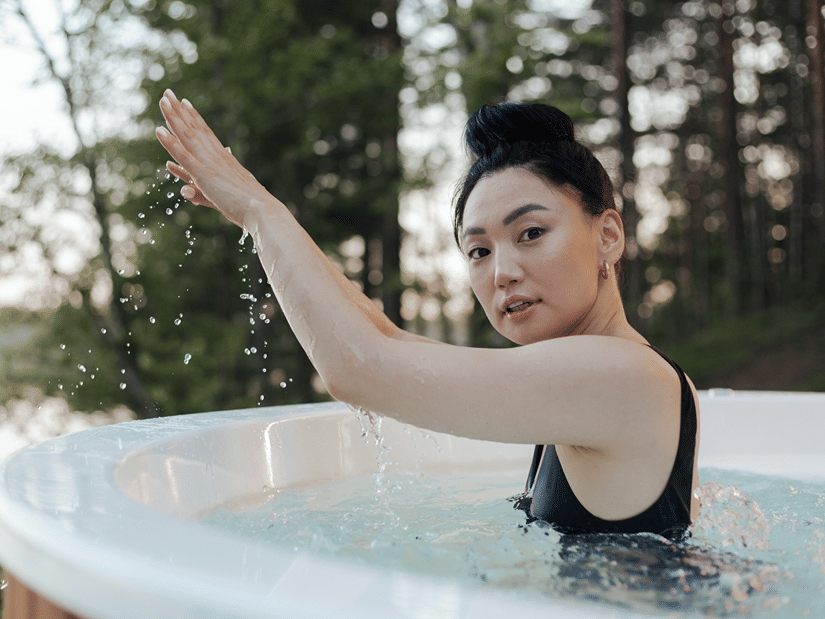 A woman in a black swimsuit splashing water in an outdoor plunge tub with pine trees behind her