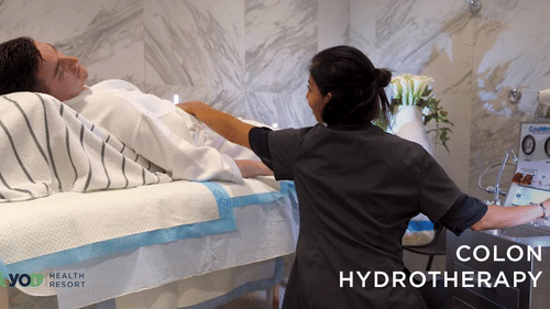 A professional in scrubs attends to a person lying on a white examination table, with equipment visible on the right, for colon hydrotherapy at YO1 Longevity & Health Resorts