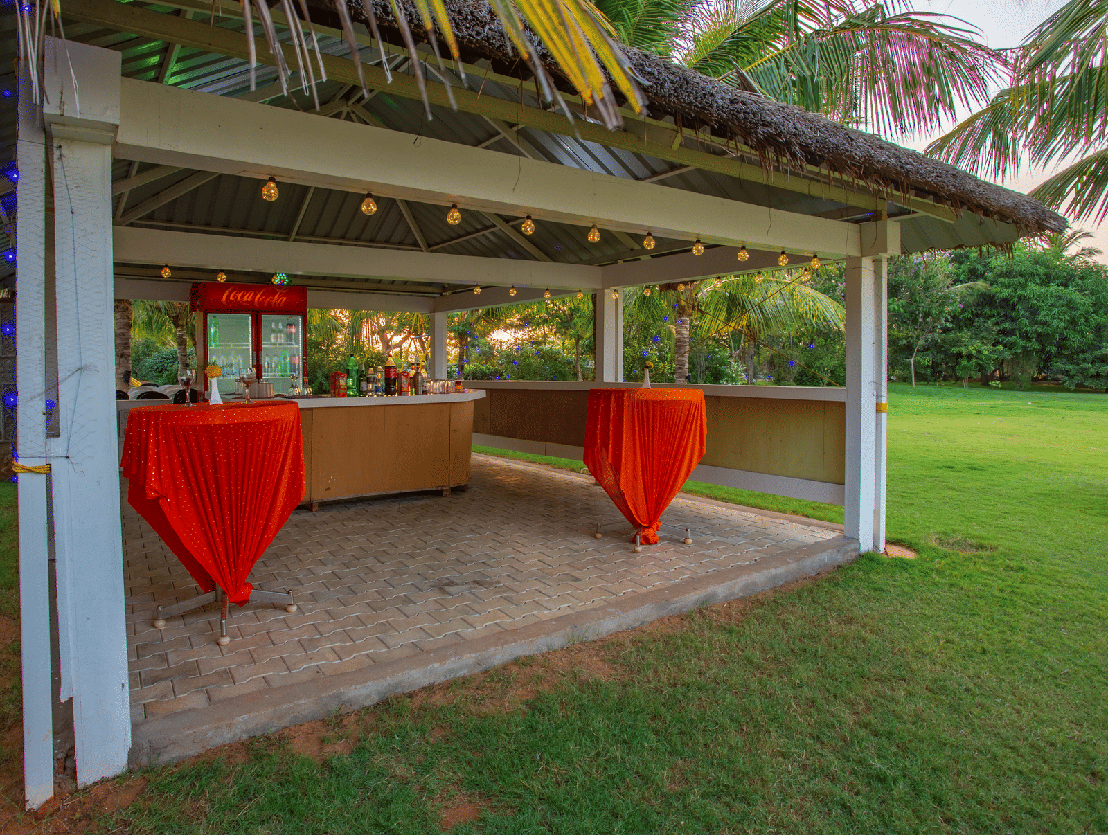 A charming outdoor bar or serving area with a thatched roof, red bar stools, and a wooden counter, set on a grassy lawn - Grande Bay Resort & Spa, Mamallapuram