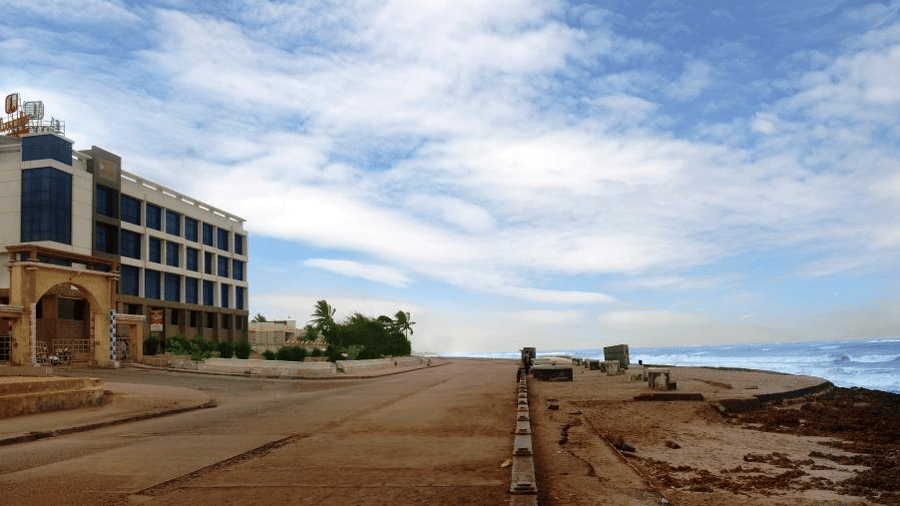 A contemporary hotel building, possibly Lords Inn Porbandar, is situated near a beach with people walking along the shore under a partly cloudy sky.
