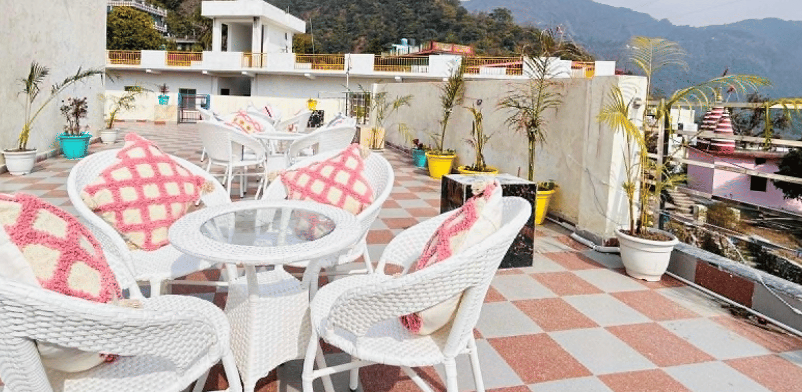 An outdoor rooftop terrace with white wicker tables and chairs featuring a red and white checkered pattern, set against a background of green hills | Perfectstayz Value Hills