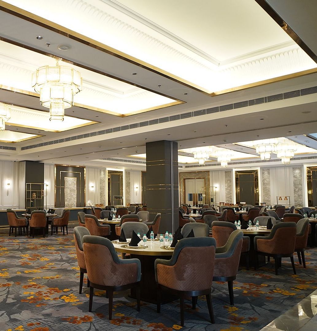 An elegant banquet hall at Nirvana Luxury Hotel, Ludhiana, featuring round tables with plush chairs on a patterned carpet, illuminated by multiple tiered chandeliers and recessed lighting.