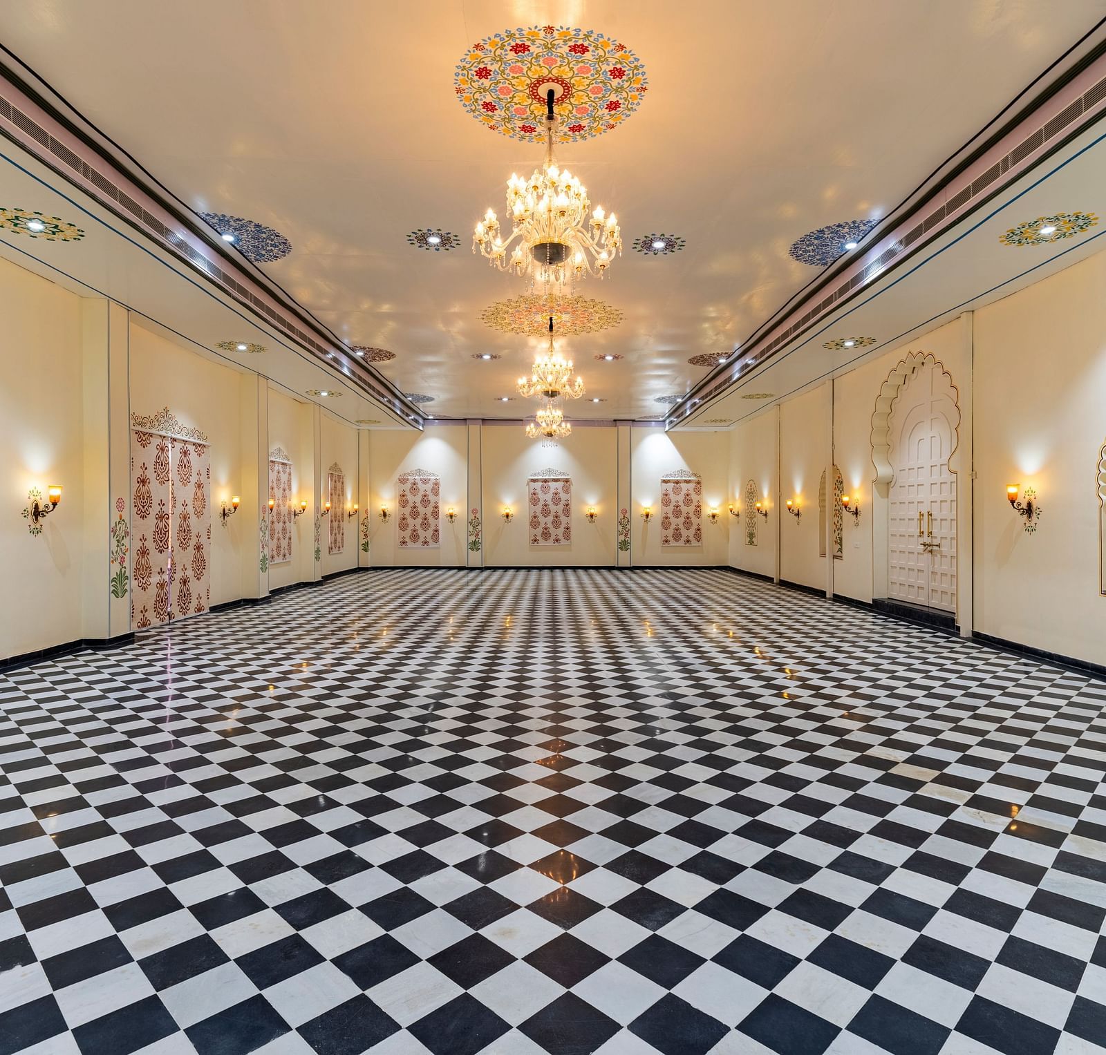Long hallway with black-and-white checkered flooring, ornate golden chandeliers, and framed artwork on cream walls.