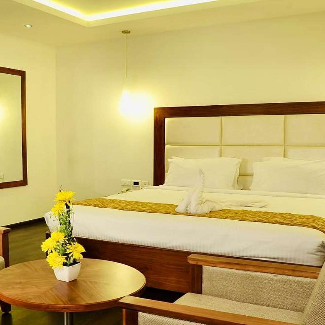 A close up view of well-lit Suite at Dhanalakshmi Srinivasan Hotels, Perambalur featuring a king size bed, two armchairs, a small table, and a work desk with a chair.