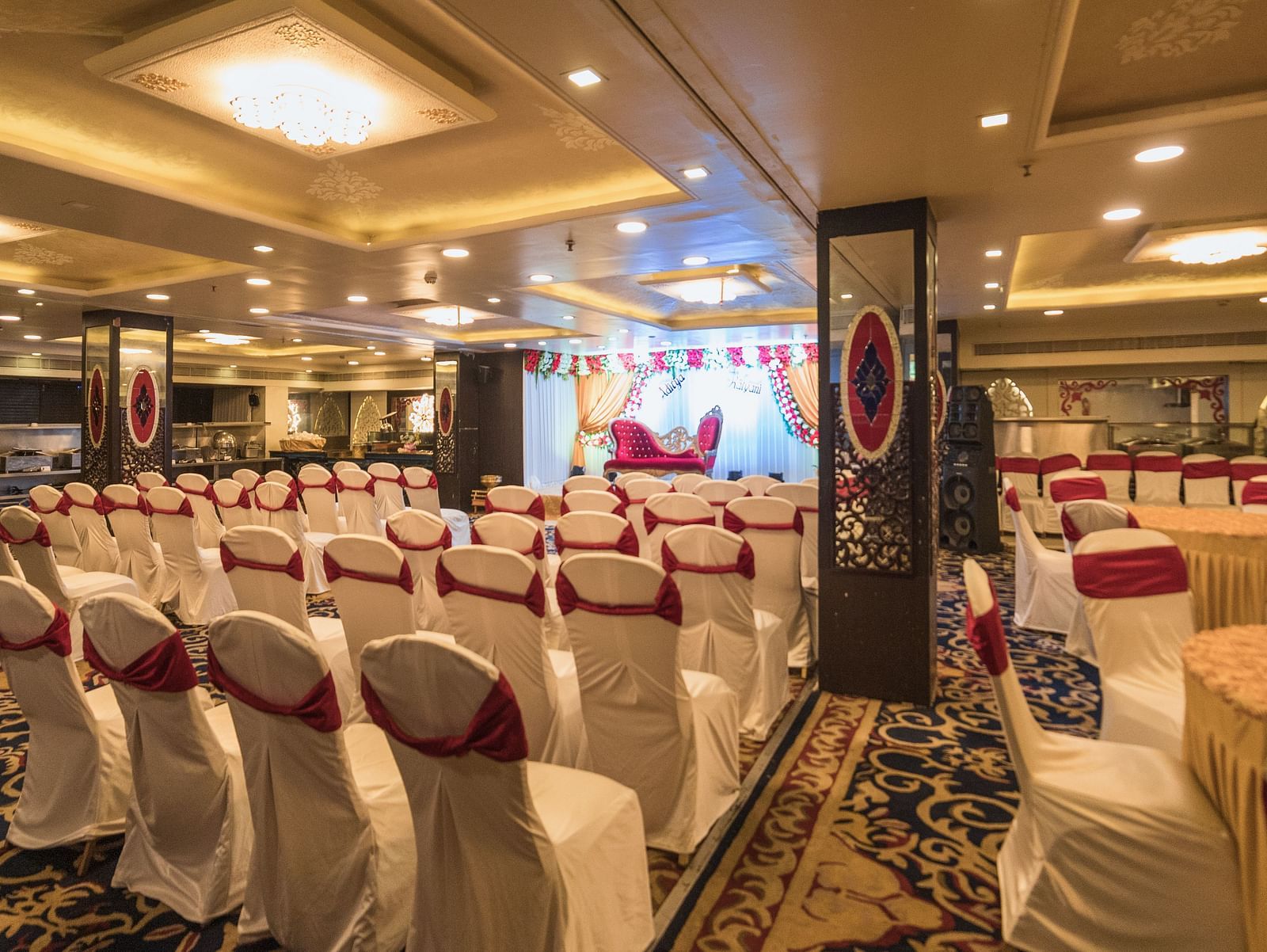 Maurya featuring rows of chairs with white covers and red sashes facing a stage area with draped fabric and lighting.