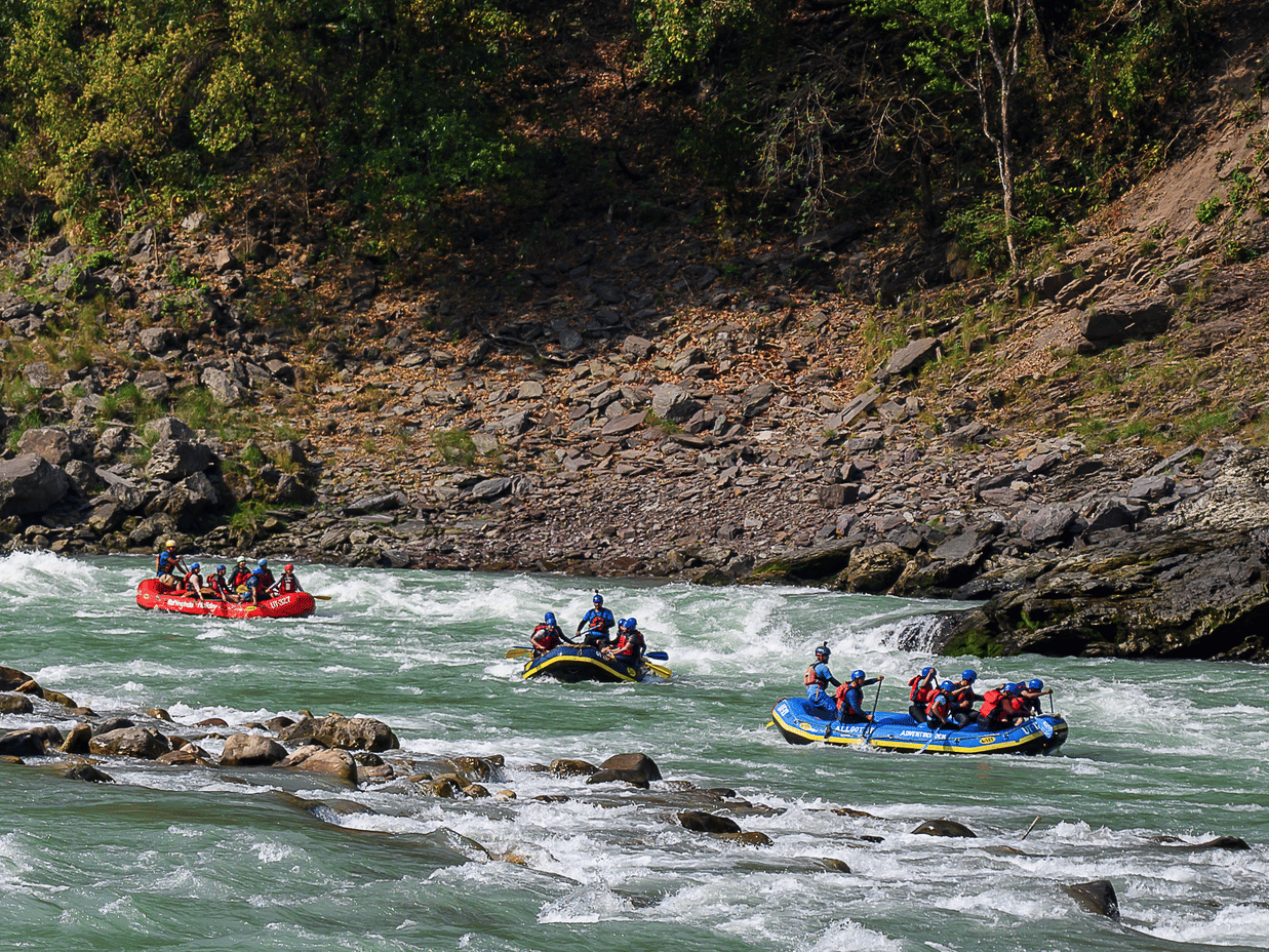 Three blue inflatable rafts filled with people navigating the white water rapids of the river past rocky banks capturing the exciting adventure activities available to guests visiting this riverside retreat