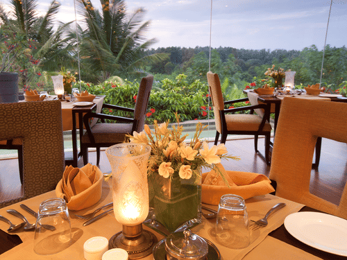 Candle-lit dining tables with plates, cutlery, napkins and floral centrepieces placed indoors, facing large glass windows that overlook greenery and palm trees at The Serai Chikmagalur.