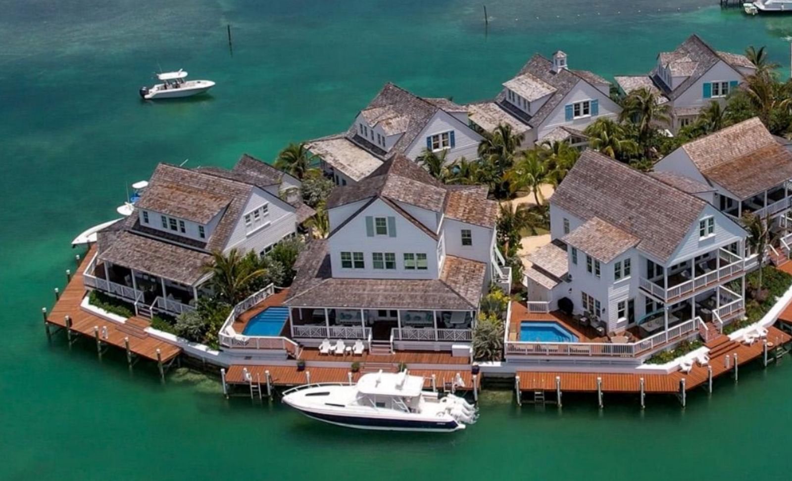 Multiple white waterfront homes with docks, some with pools, on a lush green island in turquoise water, with boats moored and another cruising near Abaco Inn.