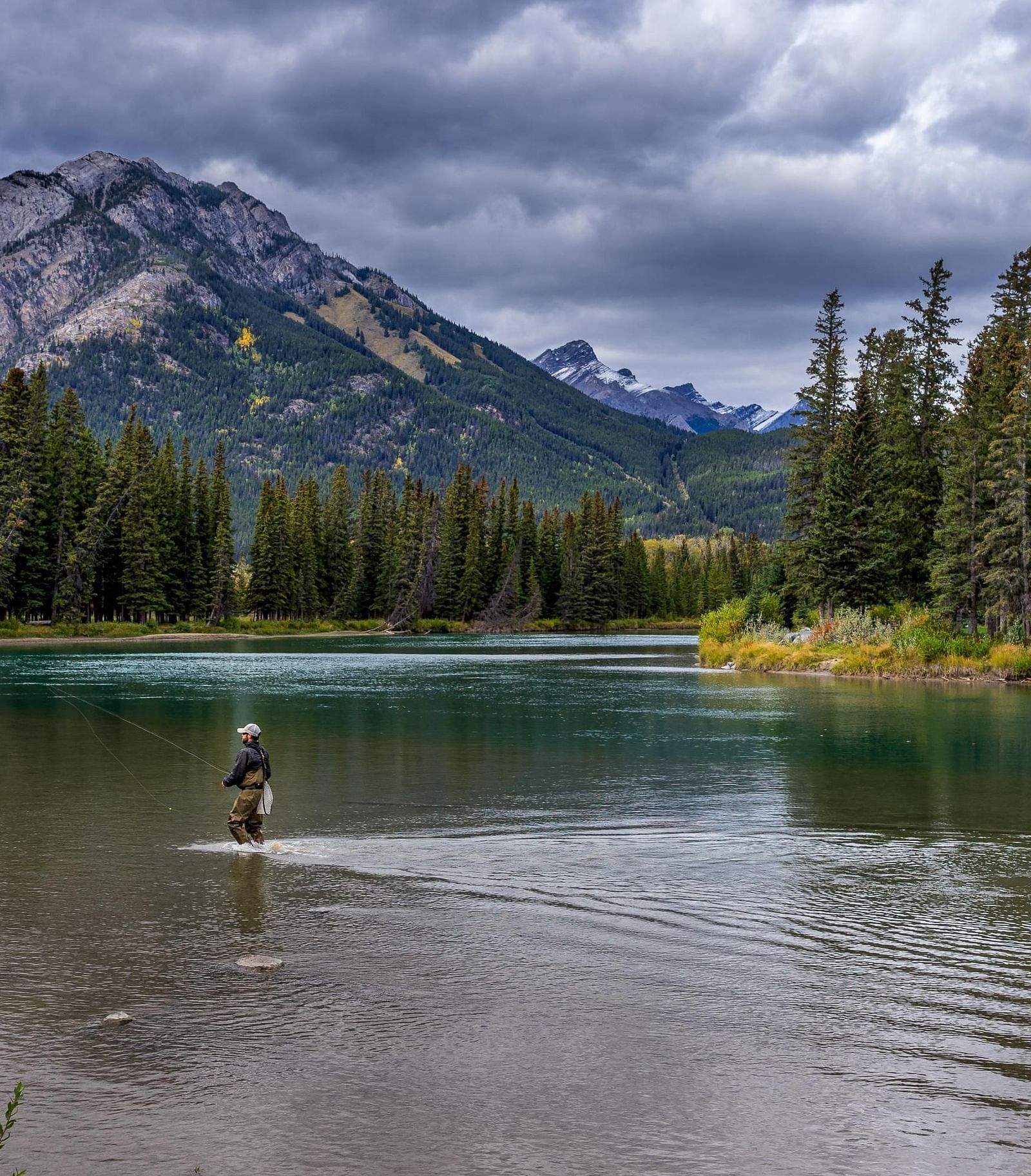 A lone person fishing in a river surrounded by greenery and a mountain.