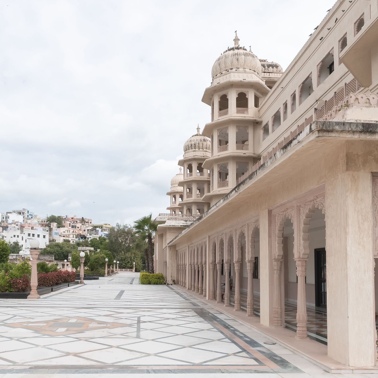 An elegant exterior view of Shri Vallabh Vilas Lords Plaza Nathdwara, showcasing intricately carved arches, domed pavilions, and a spacious marble courtyard lined with decorative lamp posts and greenery.