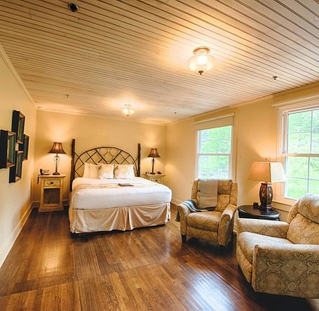 full view image of a room with wide windows featuring a cozy double bed and comfortable cushioned armchairs in front of it - Historic Tapoco Lodge
