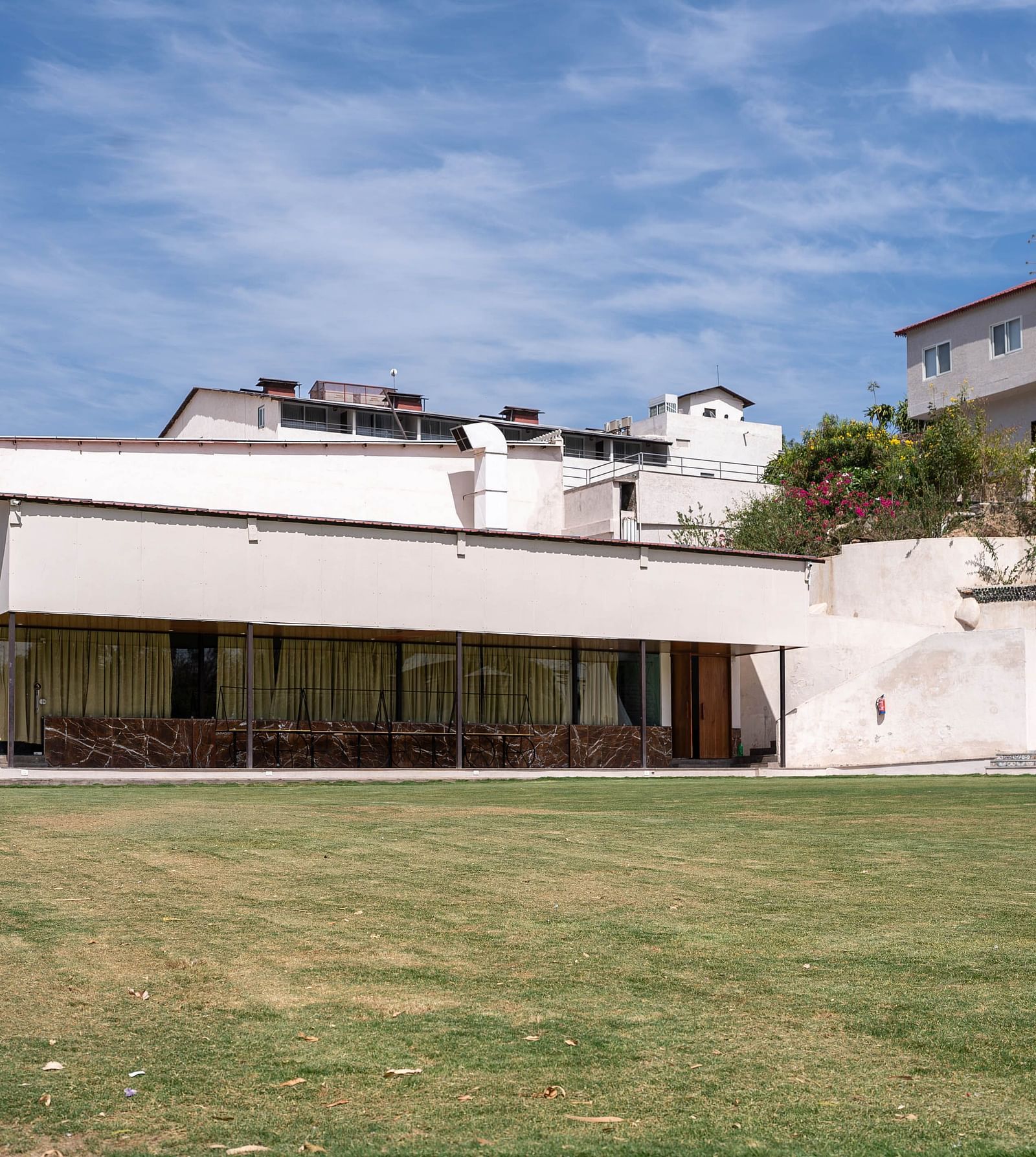 A facade of the white building of Bamboo Saa Resort & Spa, Udaipur with a flat roofline and a blue door set against a blue sky with white clouds.