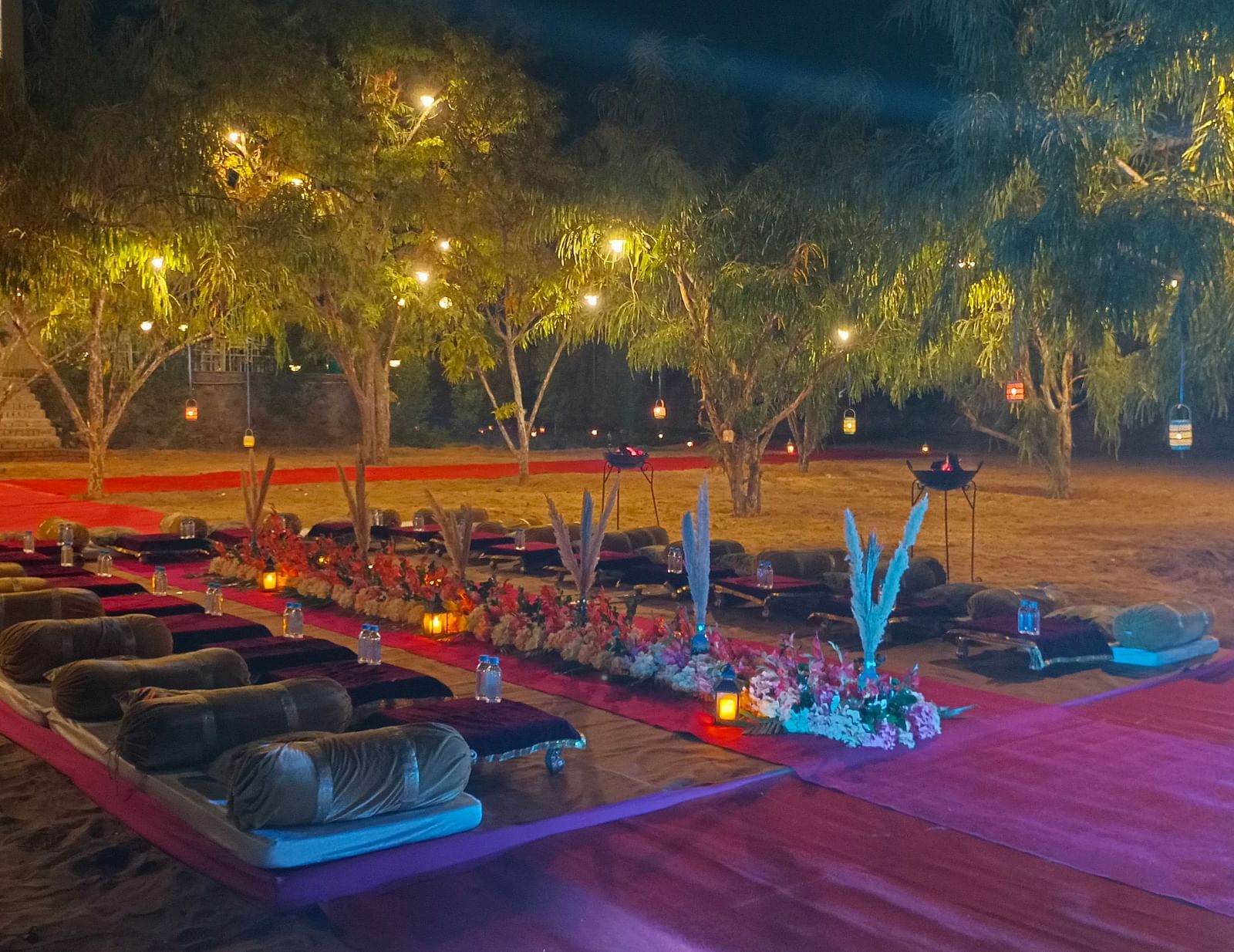 An outdoor area at night at the Bamboo Saa Mulberry Resort, Pushkar, with seating arrangement on the ground illuminated by string lights.