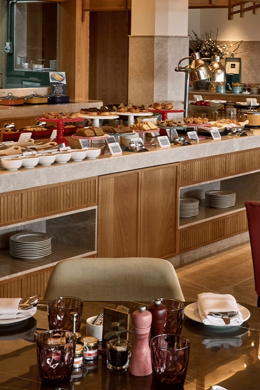 Bright indoor restaurant with a long buffet counter displaying food, and dining tables with red upholstered chairs.