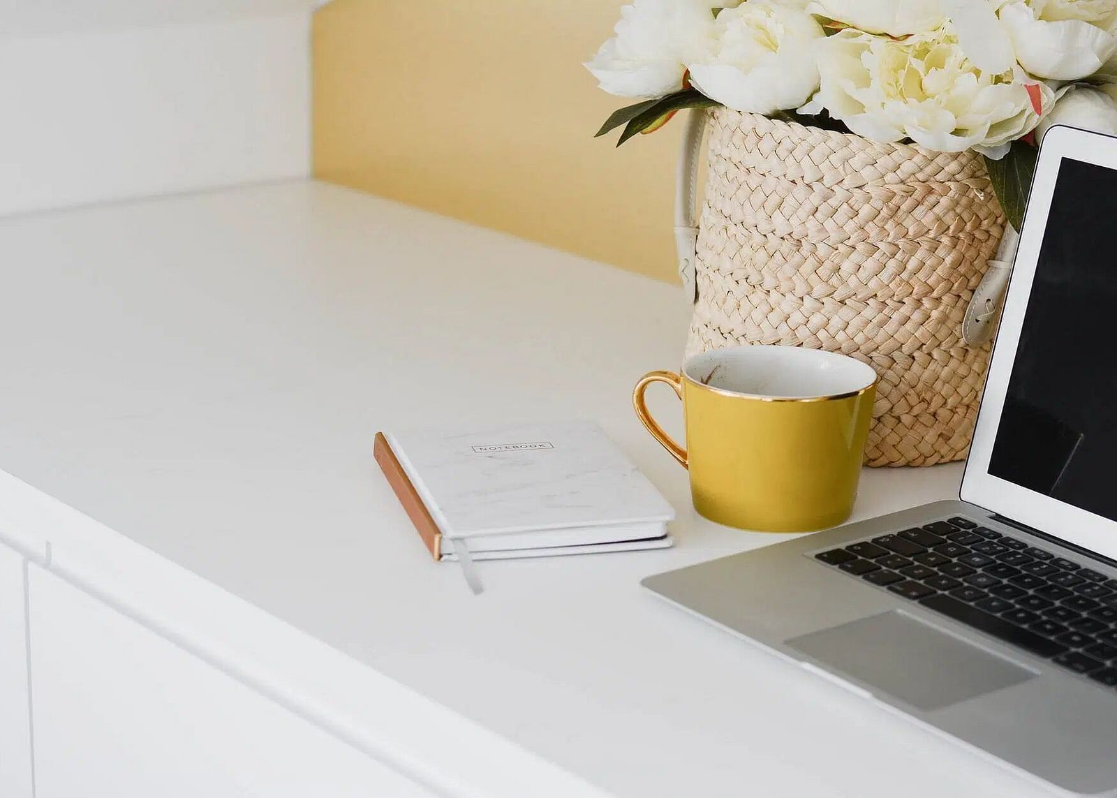 Clean, workspace with a laptop, notebook, mug, and flowers in a woven basket. 