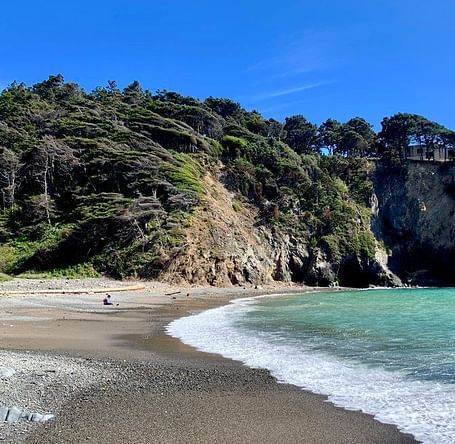 A curved shoreline meets a cliffside covered in vegetation, with a structure perched atop the rocks under a clear sky.