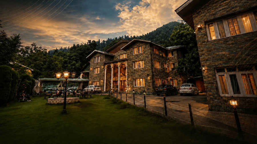 Exterior view of Hotel Outlook, Pahalgam, showing stone buildings next to a lawn, illuminated under a cloudy sky with hills in the background.