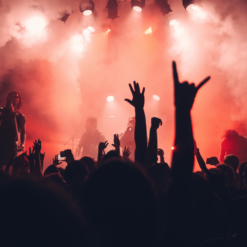 Crowd at a concert with hands raised in the air, lit by red stage lights and fog.