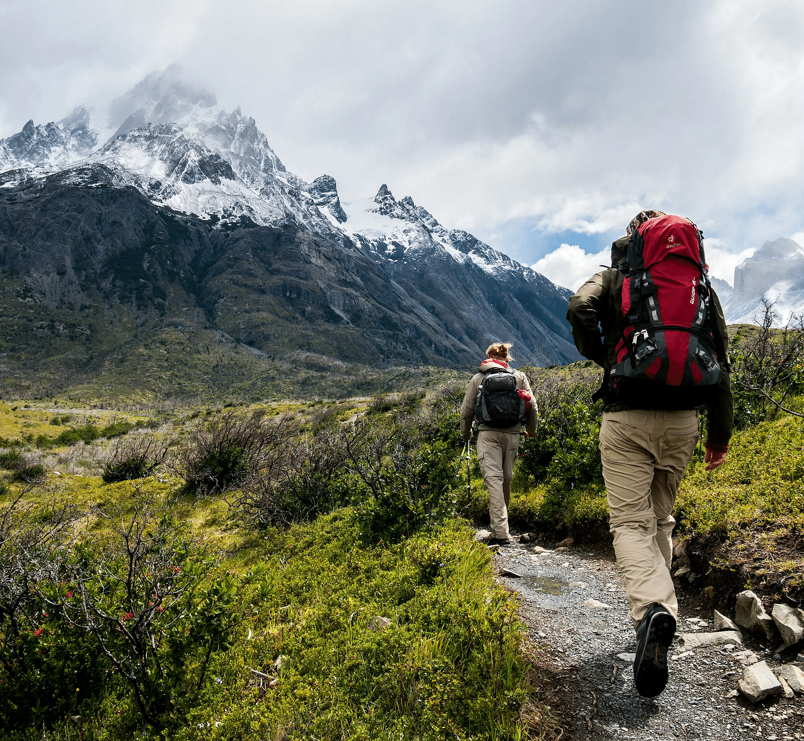 Two people trekking through a hilly region with a well packed rucksack on their backs and towering mountains in front of them.
