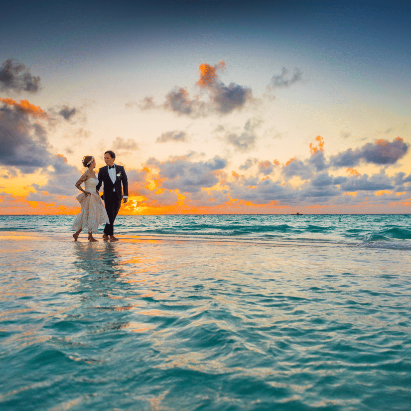 A wedding picture of a couple walking on the shallow seashore in the evening with a sunset view in the backdrop.