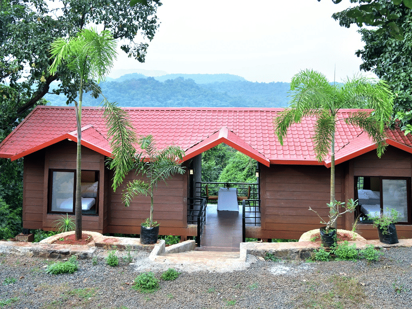 Twin red-roofed wooden cabins in a forest setting with a mountain backdrop at Nature Trails Kundalika