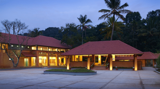 The facade of Abad Green Forest, Thekkady, featuring a single-storey structure with red sloping roofs, wide entrances, and a spacious open courtyard, set against a backdrop of lush greenery and tall palm trees.