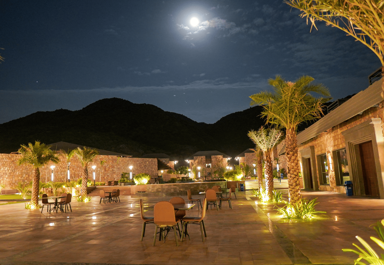 A resort courtyard is illuminated at night with warm, golden lights under a full moon. There are tables and chairs set up for al fresco dining.