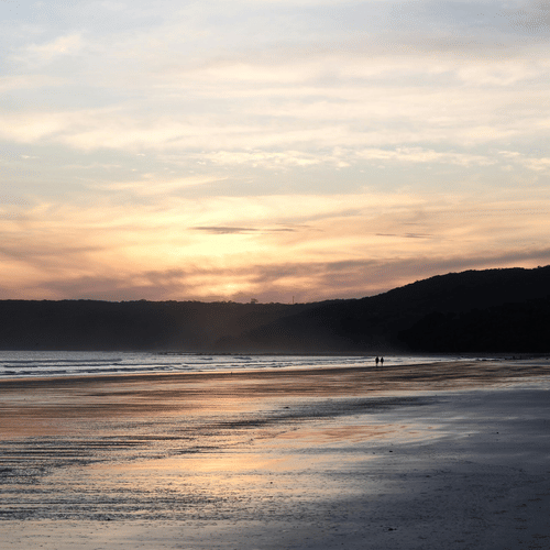 A beach while the sun is setting with the orange sky and other rocks at the back.