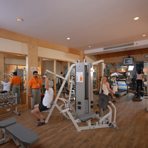 A well-equipped gym has several people using different exercise machines, with staff members in orange shirts seemingly assisting at UDS Group of Hotels.