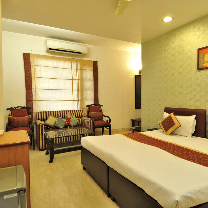 A bedroom featuring at The Cosy Hotels featuring a bed stands near a wall. Pillows and a runner rest on the bed. A sofa sits by a window. Chairs flank the sofa. A table with a top made of glass is in front of the sofa. An air conditioner hangs above the wi