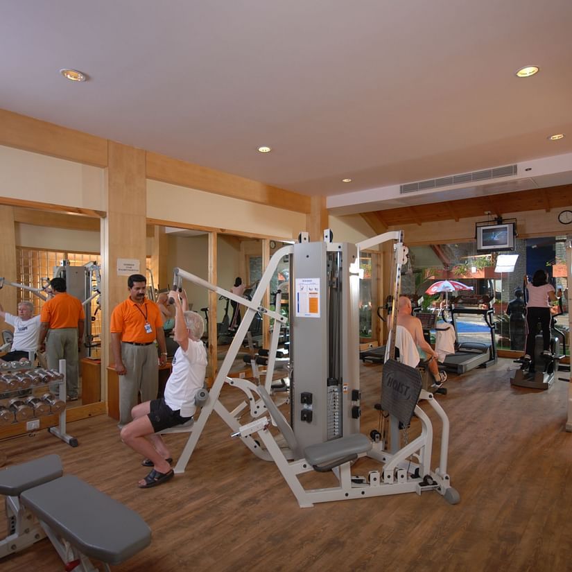 A well-equipped gym has several people using different exercise machines, with staff members in orange shirts seemingly assisting at UDS Group of Hotels.
