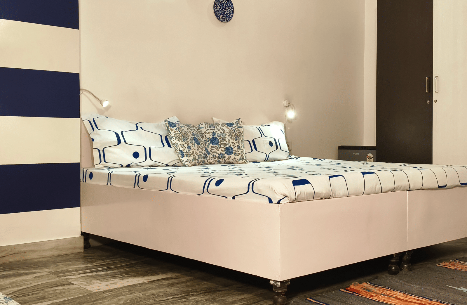 A neatly arranged bedroom with a double bed, blue accent wall, bedside lamp and rug on the floor at Jaypore360 Homestay - Best Rooms in Jaipur.