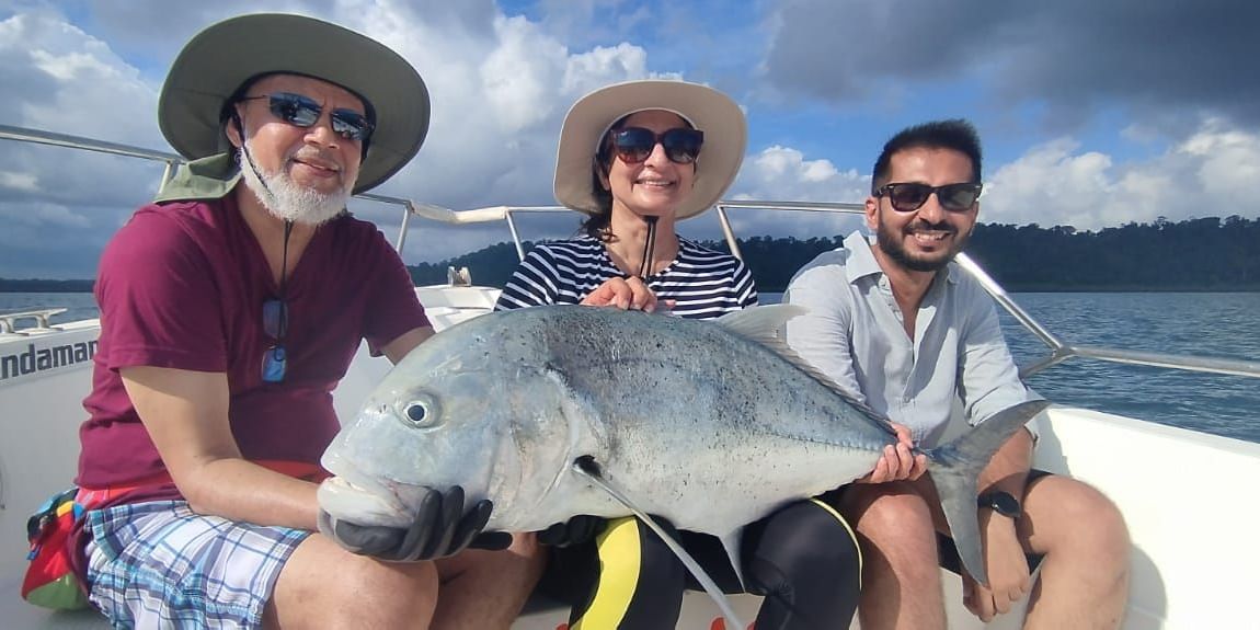 Three people sitting in a boat and holding a big fish. At the background there is a river and beyond that a land full of trees.