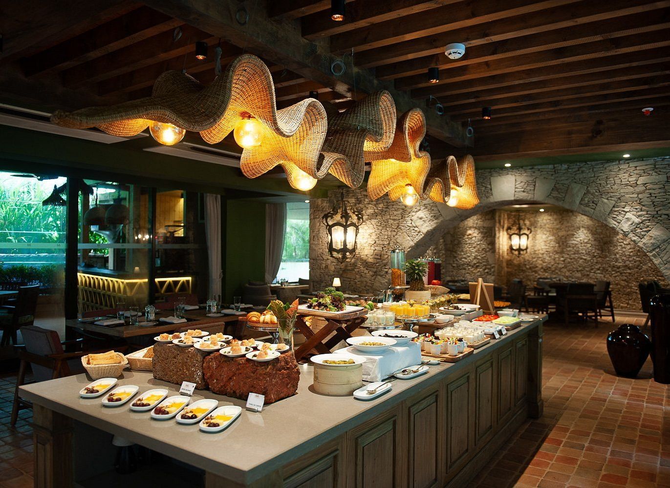 A lavish breakfast buffet spread at Mila Restaurant at The Evren, Vagator, featuring a variety of dishes on a large central island. The restaurant has a rustic yet modern ambiance with woven light fixtures, a stone archway, and a view of the outdoors.