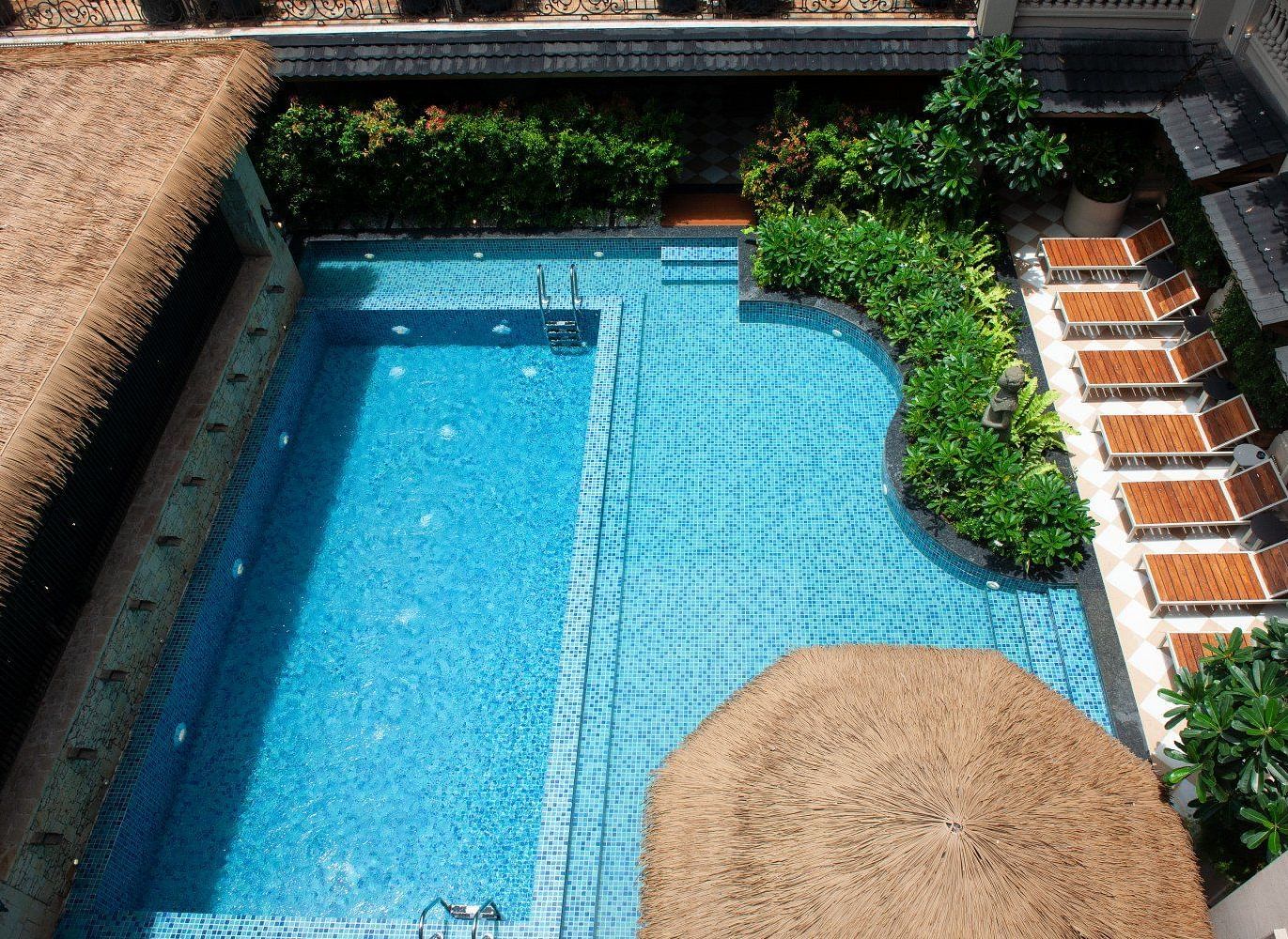 An aerial view of the outdoor swimming pool at The Evren, Vagator, featuring bright blue mosaic tiles, a built-in seating area, and a row of lounge chairs shaded by a large straw umbrella. The pool is surrounded by lush green plants and is overlooked by th