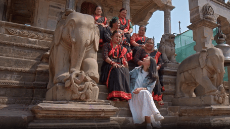 A group of girls in matching outfits sits on stone steps with elephant carvings on both sides, while a girl in modern clothes sits facing them, smiling
