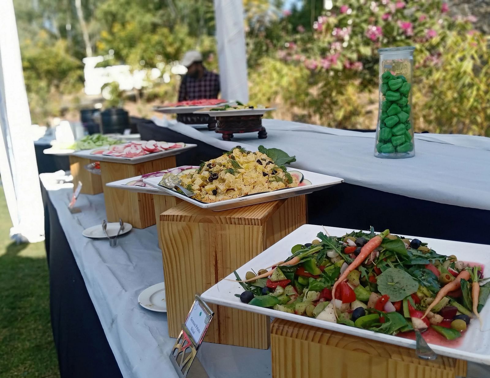 A buffet table with neatly arranged salads and food in an outdoor setting with white cloth draped on the table.