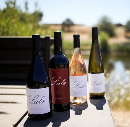Four bottles of Lula wine stand on a table outdoors, with a blurred background of greenery and a body of water.