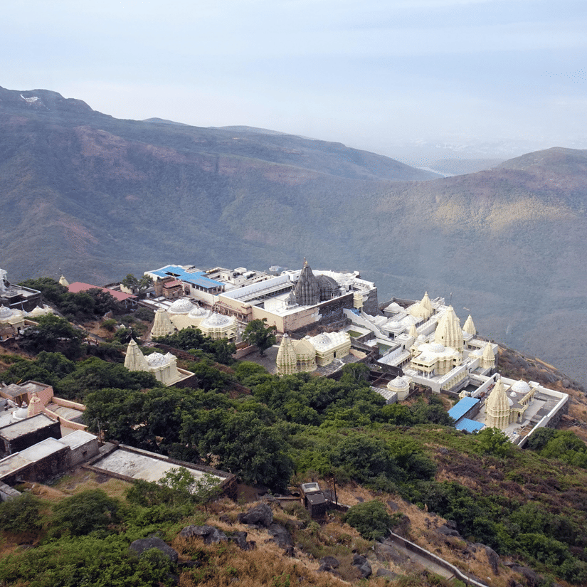Aerial view of a hilltop temple complex surrounded by lush greenery and rolling mountains, showcasing sacred architecture and scenic landscapes.