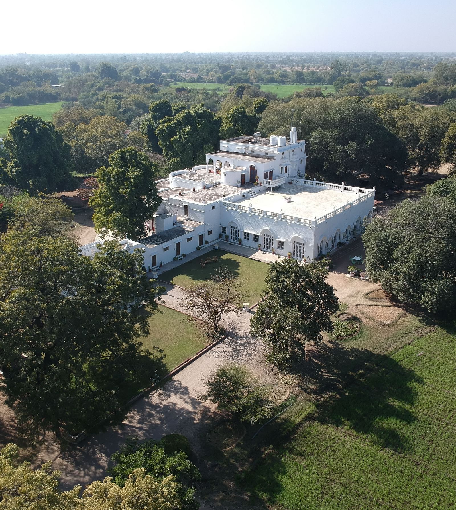 An aerial view of a white building complex with a central courtyard, surrounded by lush greenery and fields- Shahpura Bagh.