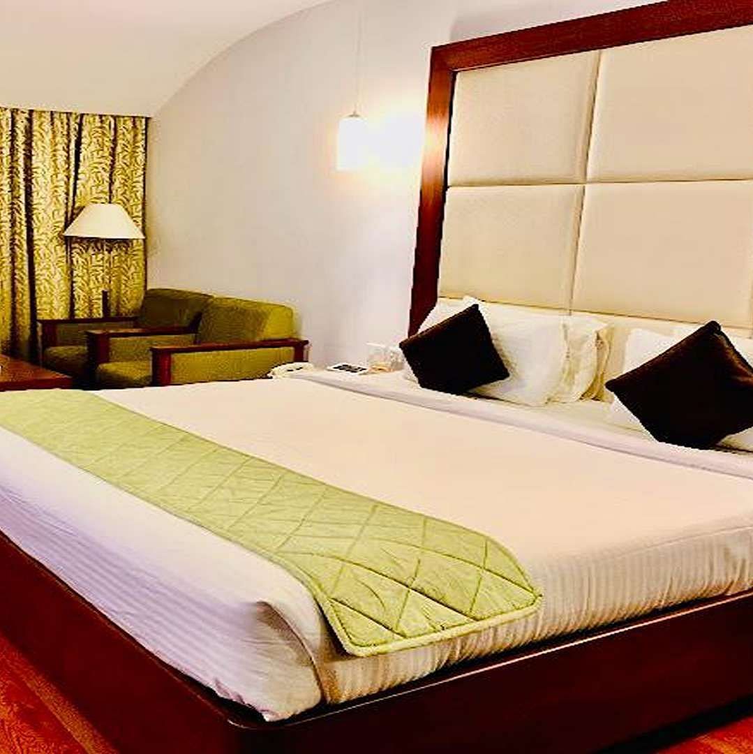 Side view of a Executive Suite at Dhanalakshmi Srinivasan Hotels, Perambalur featuring a bed with white linens, light wood flooring, and a mini-fridge.