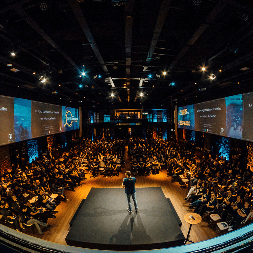 A view from the top of a speaker speaking to a crowd who are seating in a theatre-style inside an event space.