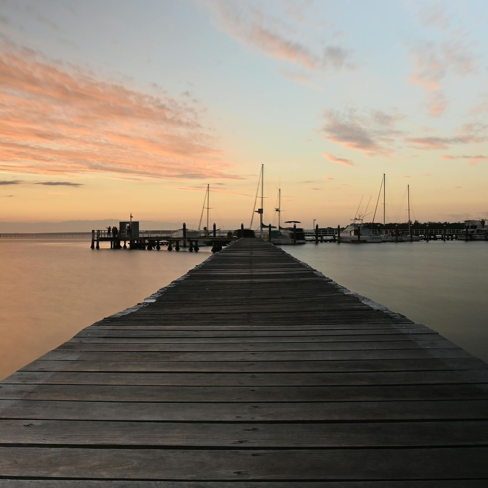 A wooden pier extending into a calm bay, with a beautiful sunset and moored sailboats in the background
