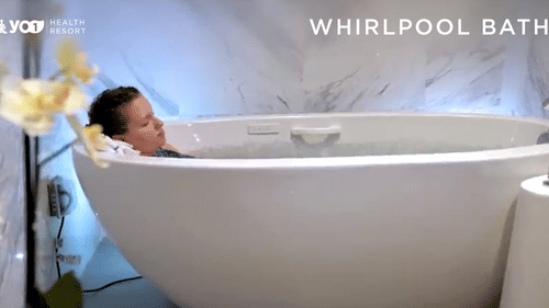 A person with short hair relaxes in a modern white whirlpool bath, with soft blue lighting and white orchid flowers blurred in the foreground at YO1 Longevity & Health Resorts
