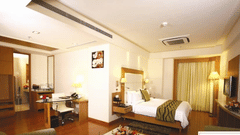  A Premium Suite at Ambrosia Sarovar Portico, Haridwar, with a bed, lounge seating, writing desk, television, and a well-lit open layout.