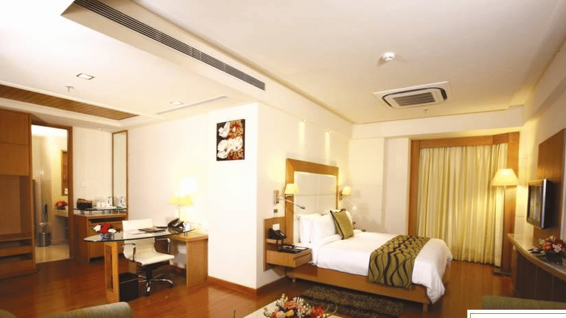  A Premium Suite at Ambrosia Sarovar Portico, Haridwar, with a bed, lounge seating, writing desk, television, and a well-lit open layout.