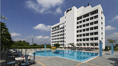 A wide-angle view of a modern hotel building behind a large, bright blue swimming pool with lounge chairs on the patio - Clarks Avadh, Lucknow