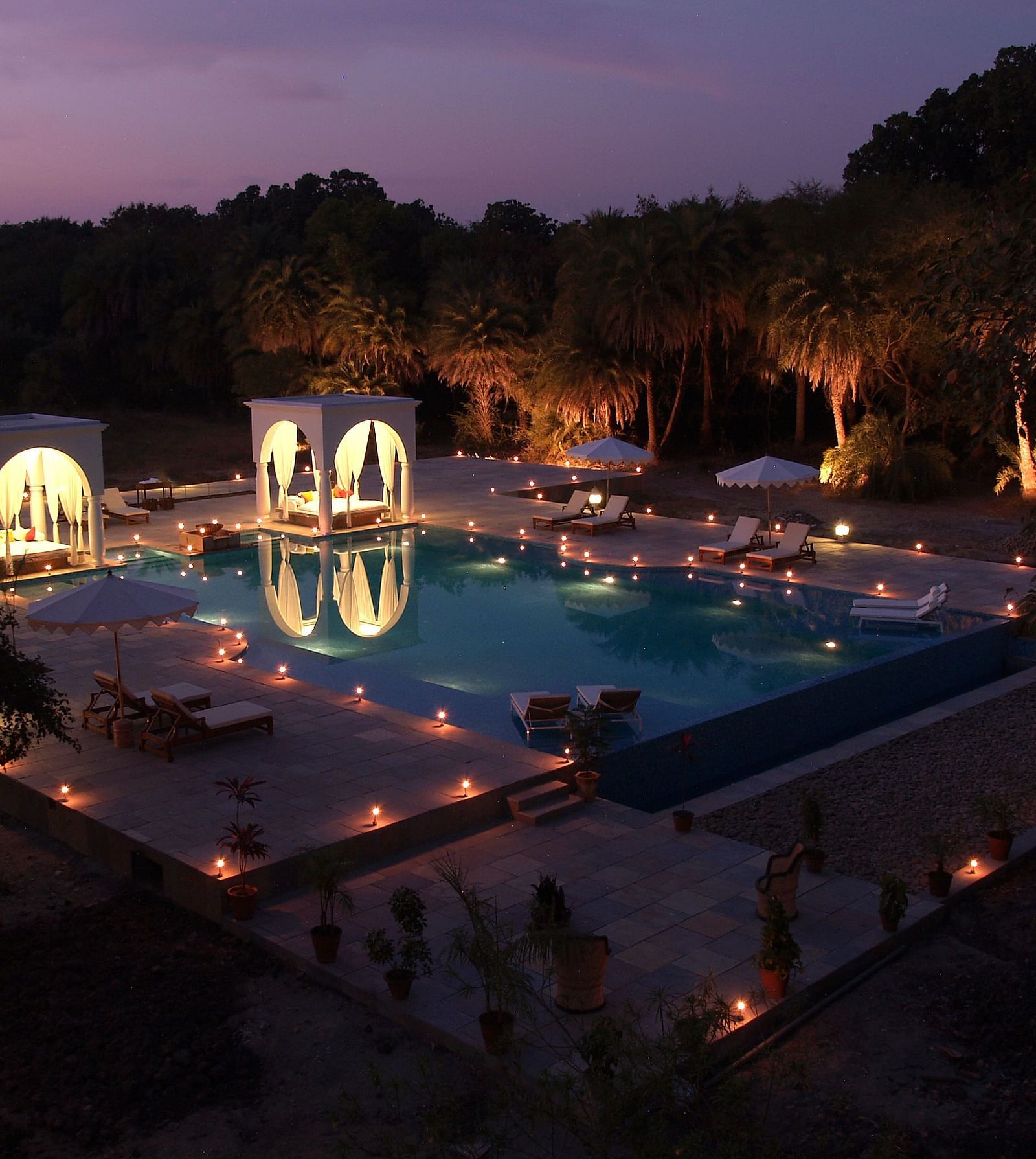 An illuminated swimming pool at night, surrounded by trees and structures with arched openings, creating a serene and inviting atmosphere- Shahpura Bagh.