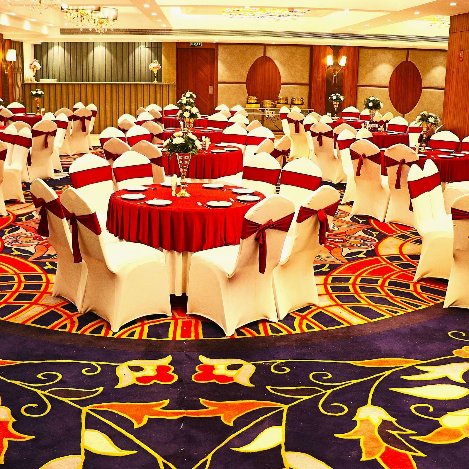 Grand banquet hall with round tables dressed in white and red covers, ornate carpet, and ambient ceiling lighting.