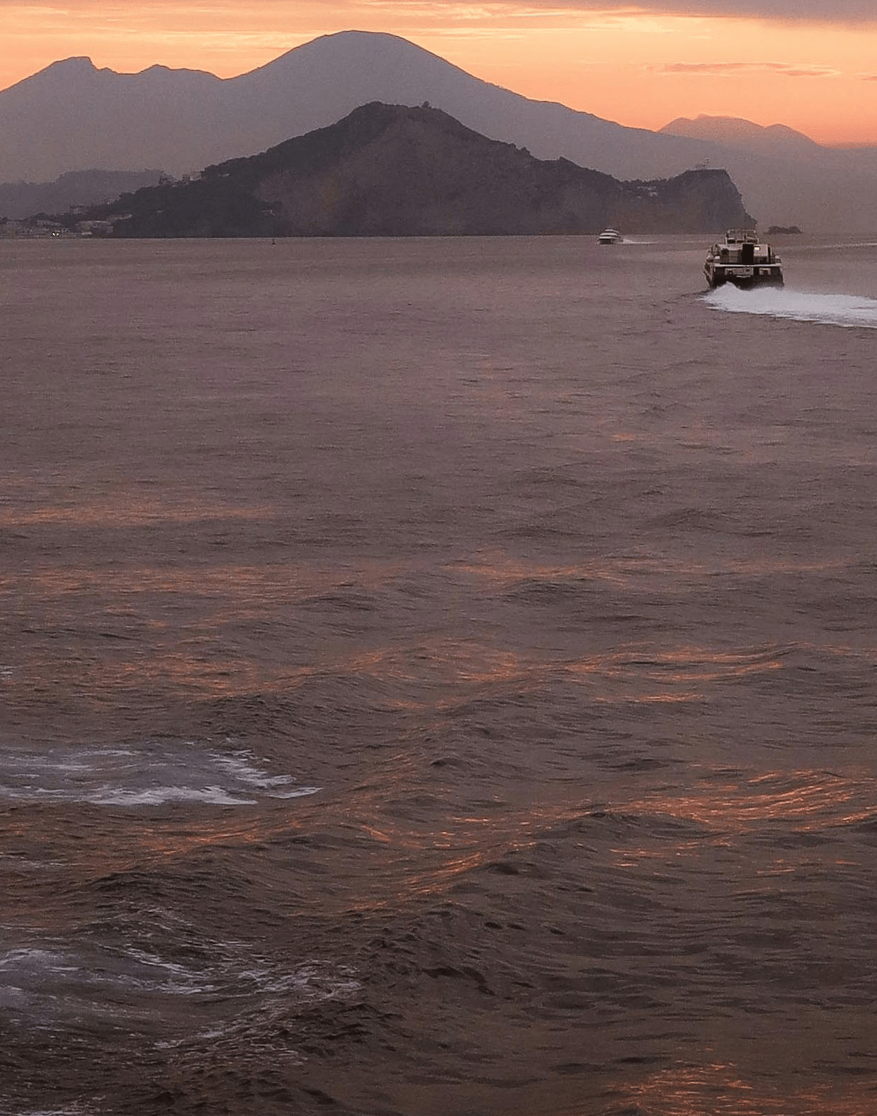 A serene sunset view over the sea, featuring silhouetted mountains in the background and soft, twilight clouds.